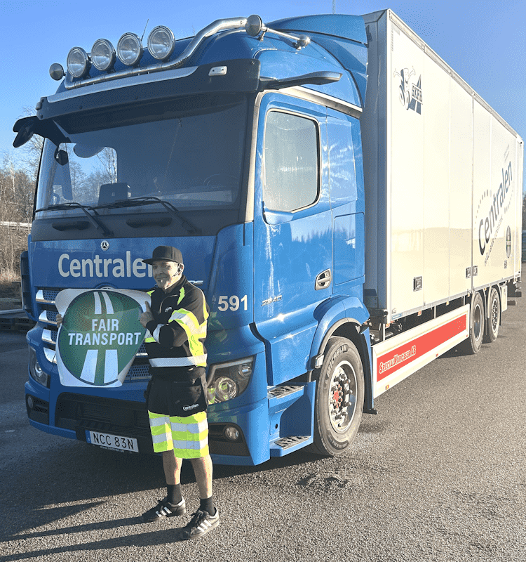 En chaufför i reflexkläder står framför en blå lastbil från Centralen och håller upp en skylt med texten "Fair Transport", vilket symboliserar företagets engagemang för en hållbar transportsektor. Lastbilen är parkerad i solljus och märkt med Centralens logotyp på sidan av släpet.